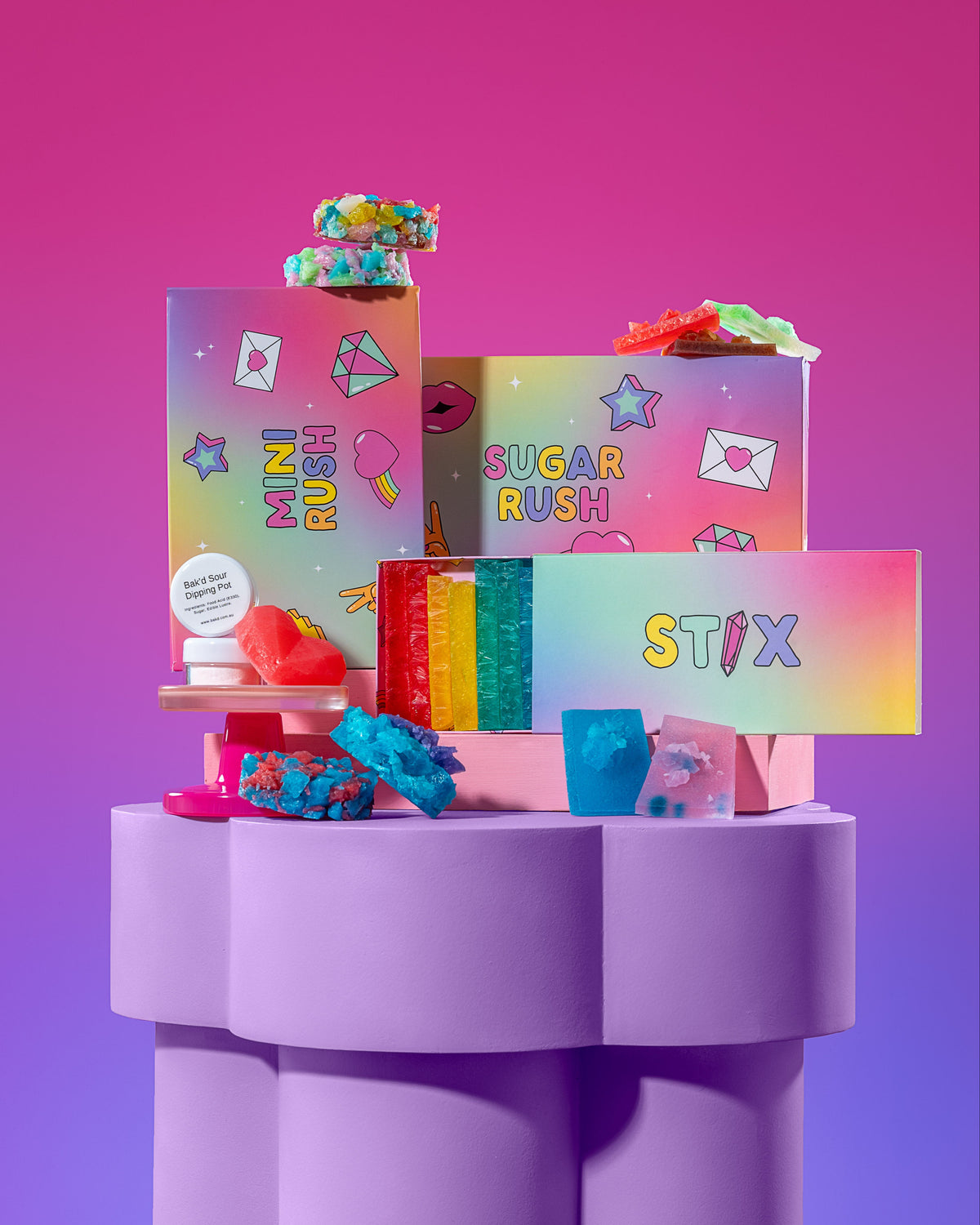 A colorful assortment of branded candy and snack boxes arranged on a lavender pedestal against a pink-to-purple gradient background. Visible text includes STIX, SUGAR RUSH, and other playful product names.