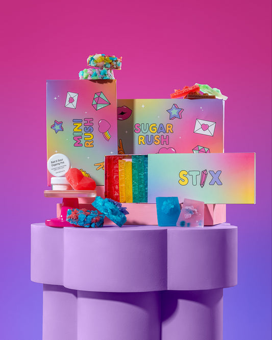 A colorful assortment of branded candy and snack boxes arranged on a lavender pedestal against a pink-to-purple gradient background. Visible text includes STIX, SUGAR RUSH, and other playful product names.