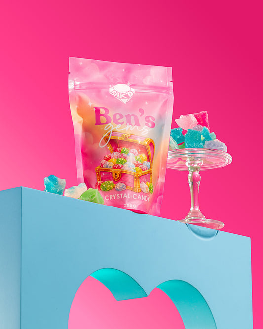 A pink stand-up pouch of Ben's Gems Crystal Candy stands on a light blue pedestal against a bright pink background, with colorful gummy candies and a glass candy stand beside it.