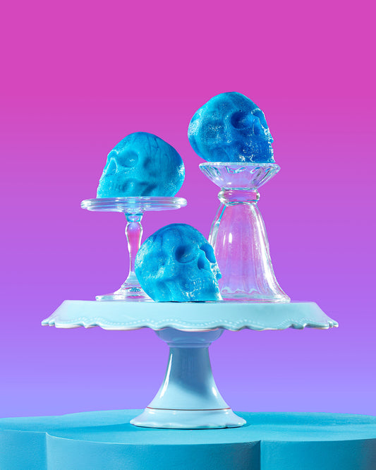 Three blue skull-shaped objects displayed on clear glass stands atop a white pedestal, set against a bright pink-to-purple gradient background.